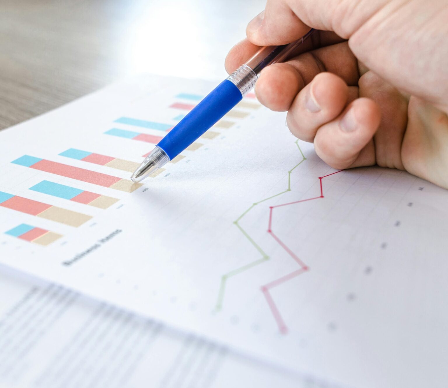 A close-up of a hand with a pen analyzing data on colorful bar and line charts on paper.